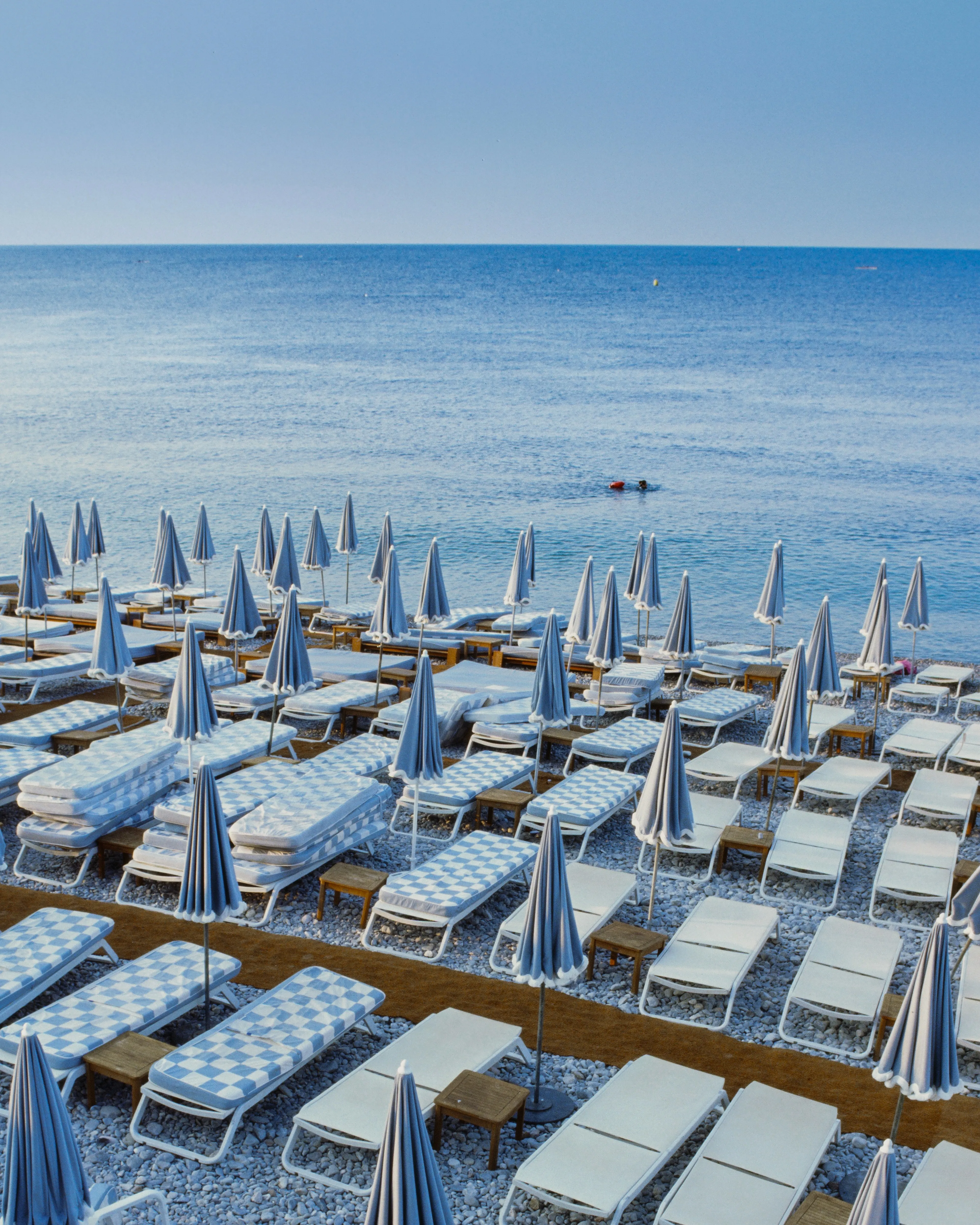 Rows of folded beach umbrellas and checkered loungers by a flat sea, one swimmer out