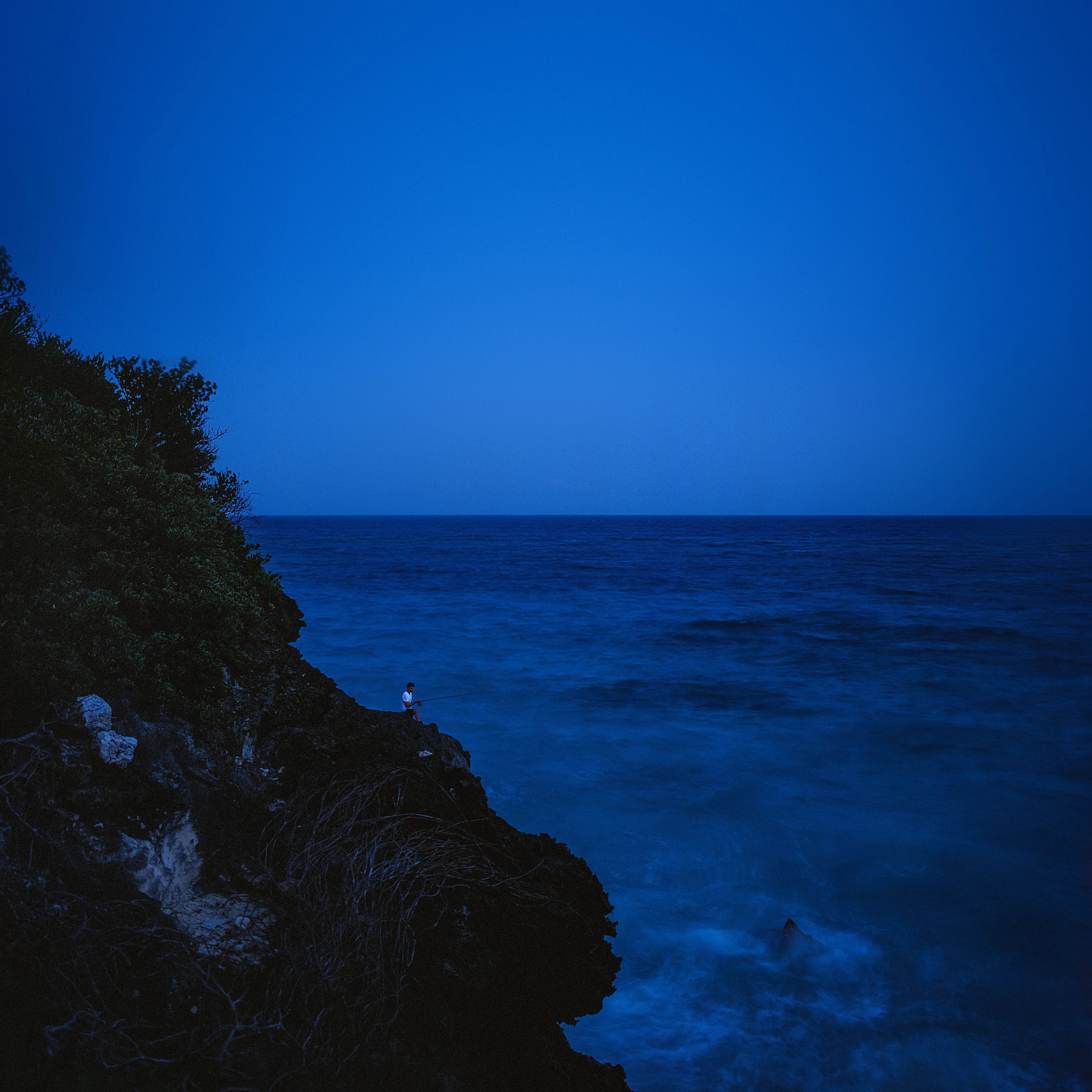 Fisherman on dark rocks at blue hour, deep blue sea