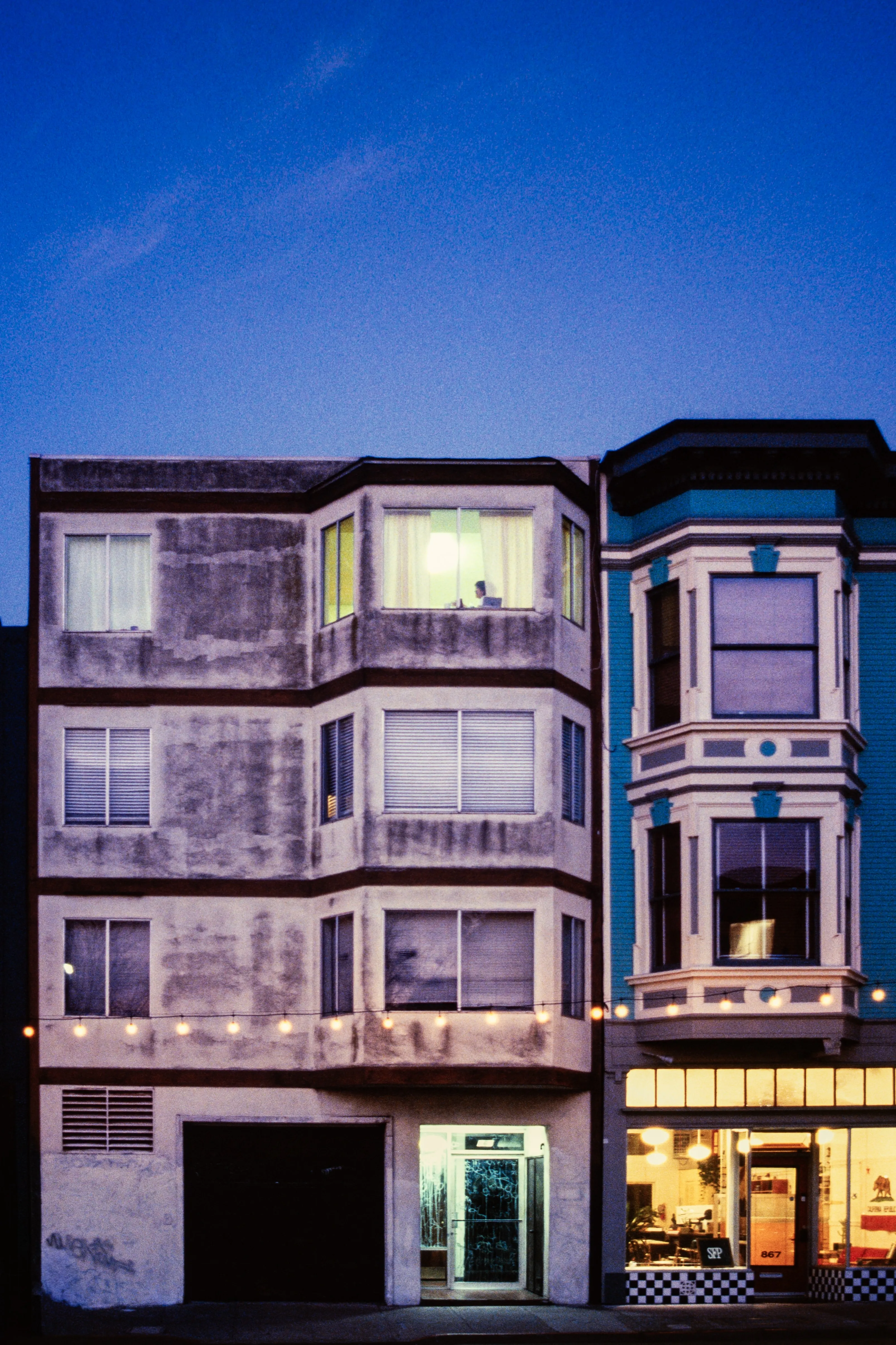 Apartment building at blue hour, a figure seated in the one lit window