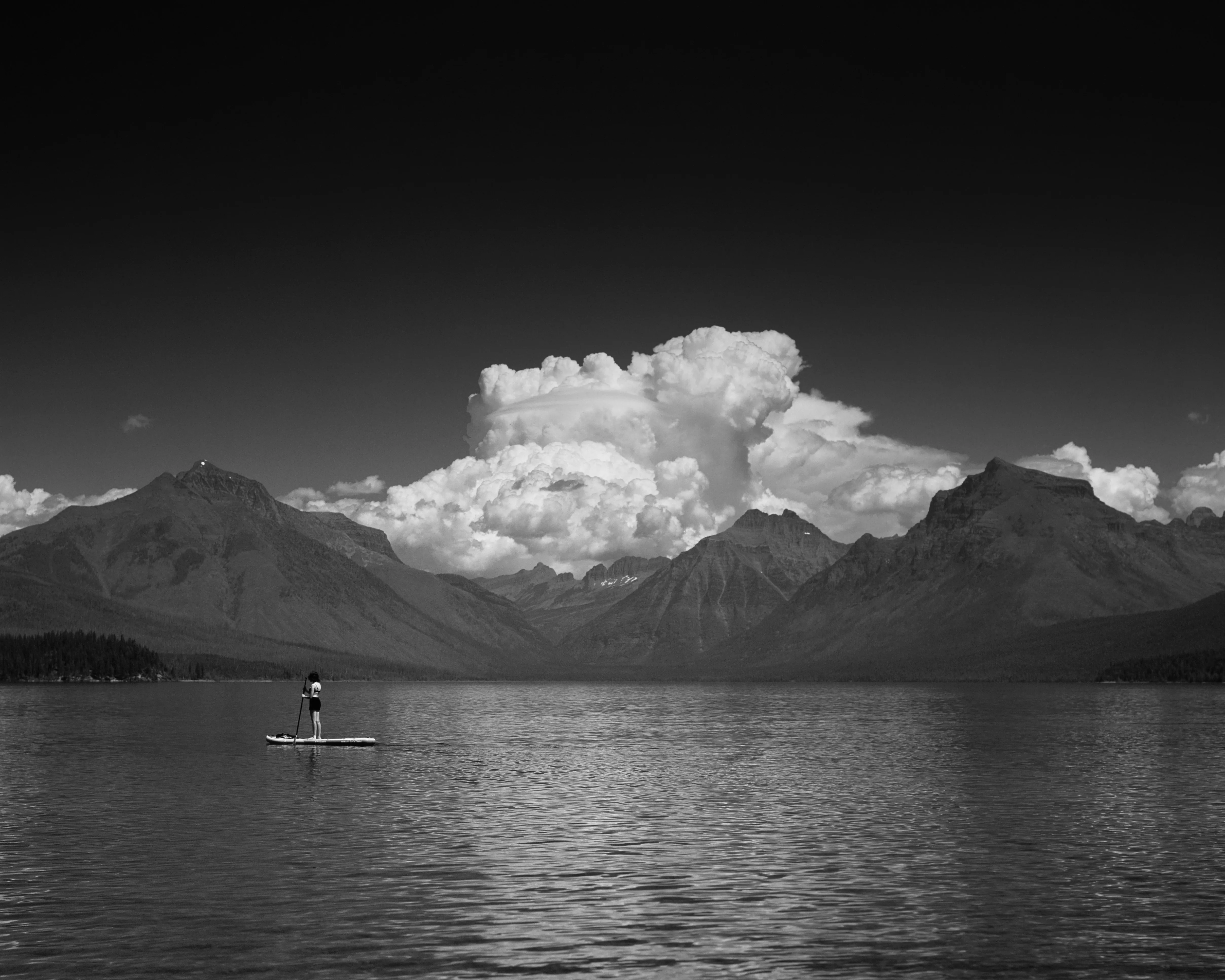 Lone paddleboarder on a vast mountain lake beneath a towering cumulus, black and white