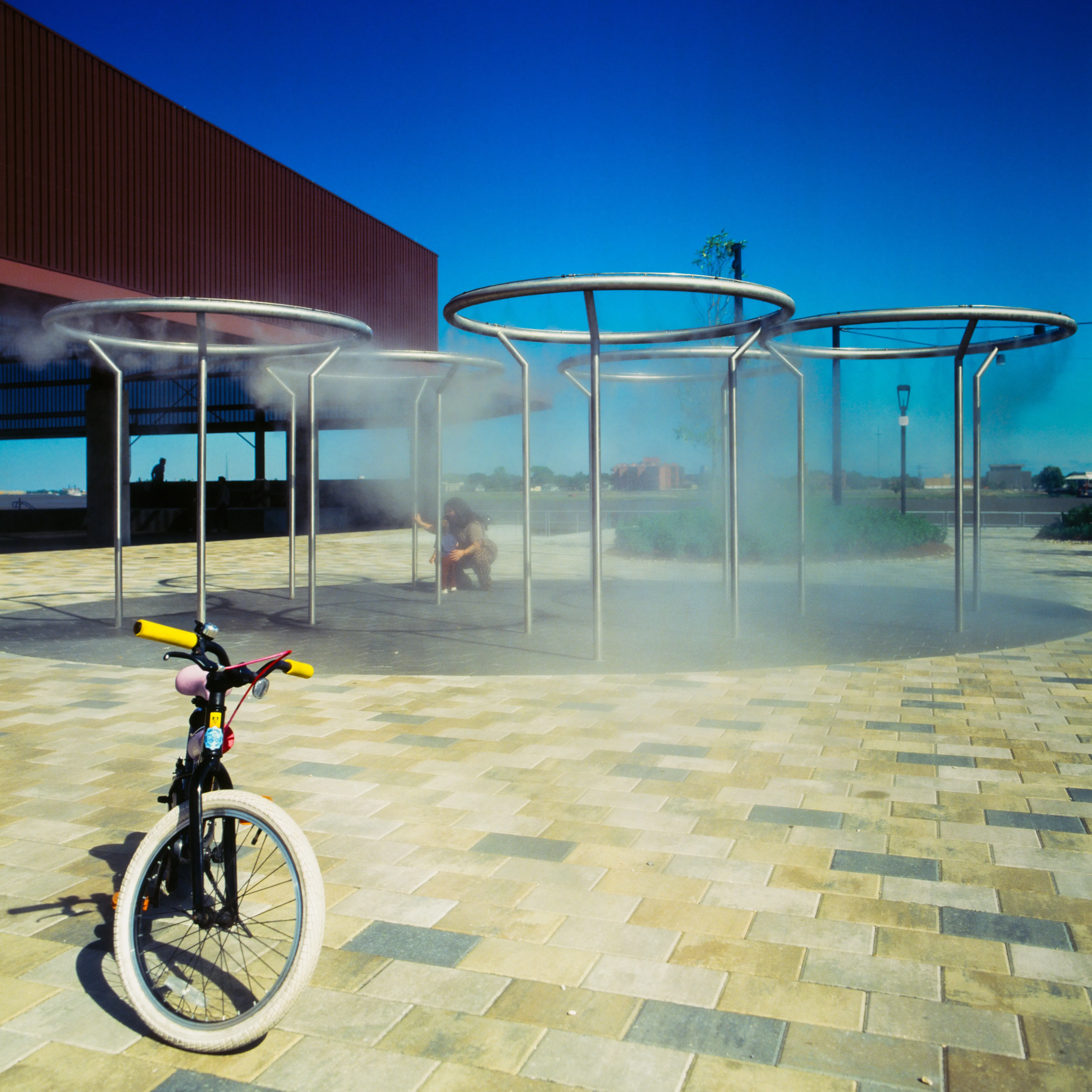 Cooling-mist canopies with figures in the steam, child's bicycle in foreground